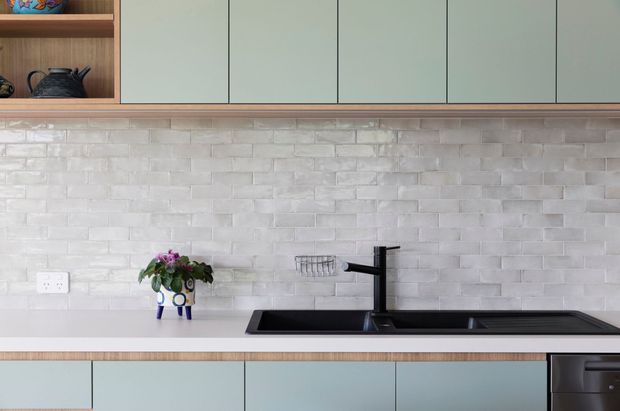 Contemporary kitchen close-up featuring muted sage-grey flat-panel cabinetry, a white textured brick-pattern splashback, a dark benchtop with an integrated double sink and black tapware, and warm timber open shelving and trim. Small decorative objects and a dishwasher are visible.