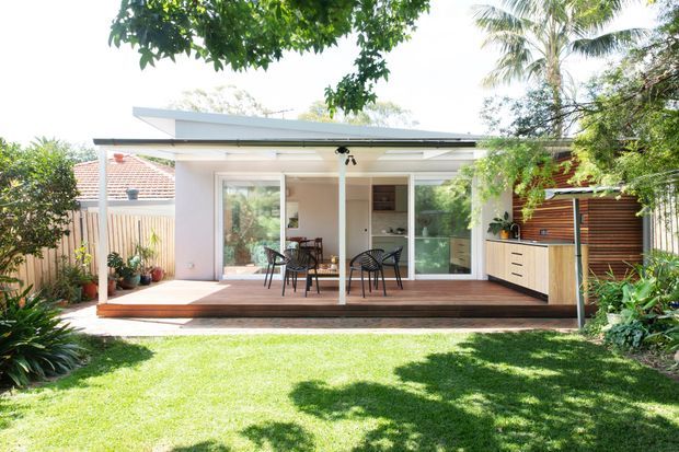A bright single-storey backyard pavilion or extension with a low-pitched roof, broad glazed sliding doors, and a covered timber deck furnished with a small dining setting. A compact outdoor kitchen or built-in cabinetry sits to the right, with timber screening, lush garden planting, and lawn in the foreground.