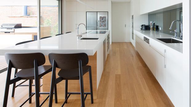 Bright contemporary kitchen with a long white island and integrated sink, dark bar stools, extensive white cabinetry, mirrored splashback, timber flooring, and large glazing opening toward an outdoor barbecue area.