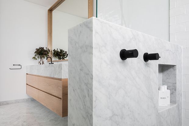 Bright contemporary bathroom featuring a freestanding marble-clad shower partition with matte black wall-mounted controls and recessed niche. A timber vanity with integrated white marble benchtop, large mirror, potted greenery, and white tiled walls are visible behind it.
