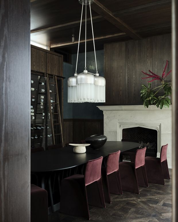 A moody, refined dining room with a long black dining table surrounded by burgundy upholstered chairs. The room features dark timber wall and ceiling panelling, a pale carved fireplace with a black herringbone firebox, a vintage-style glass pendant chandelier, built-in glass-fronted storage, and dark patterned timber flooring. A large leafy floral arrangement sits on the mantel.