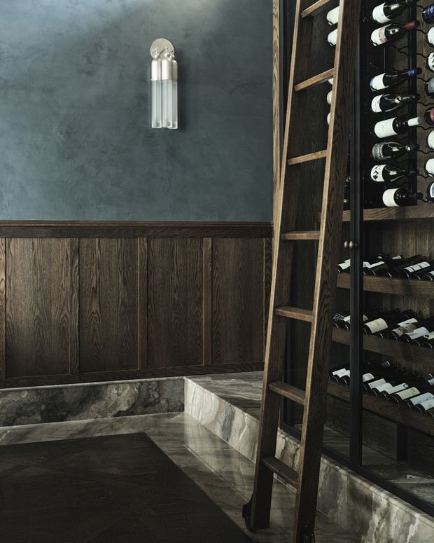 A moody, refined wine cellar featuring dark timber wall panelling and built-in wine storage filled with bottles. A timber ladder provides access to the upper racks, while a stone-clad low bench or ledge runs along the base. The space has charcoal plaster walls, dark wood joinery, polished stone surfaces, and a pair of metallic wall sconces.