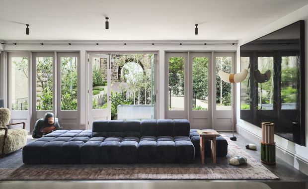 A refined contemporary living room with a large modular charcoal sectional sofa facing a dramatic black fireplace or media wall. The room has extensive grey-framed glazed doors and windows opening to a leafy garden, a pale ceiling with minimalist track lighting, a dark timber floor, a large textured rug, and sculptural side tables and decor.