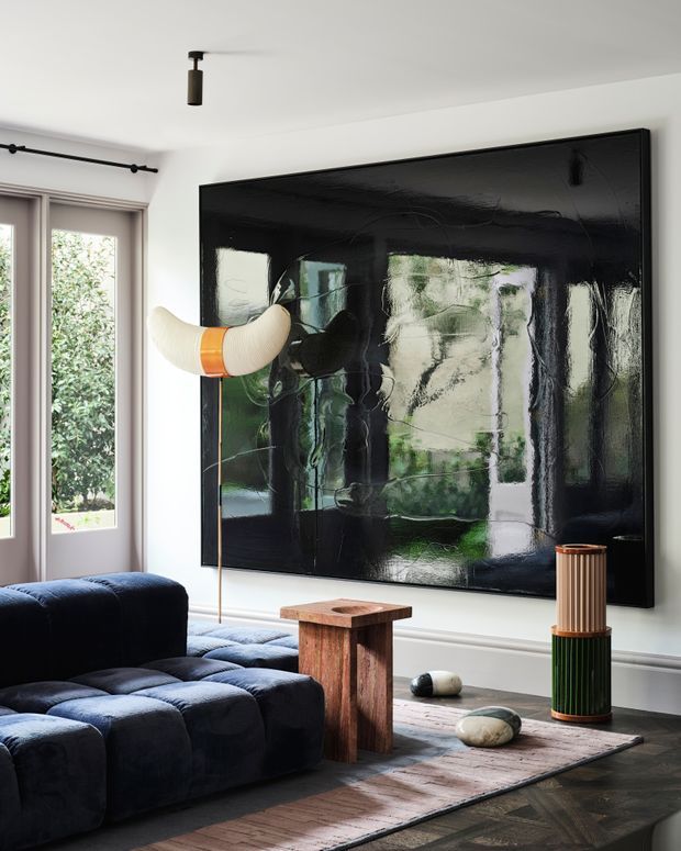 A refined contemporary living room with white walls, tall glazed doors, dark herringbone timber flooring, and a large glossy black abstract artwork. A deep navy modular sofa sits over a muted area rug, accompanied by a rustic timber side table, sculptural floor lamp, decorative stones, and a cylindrical ribbed lamp or pedestal. The room has a bold, moody yet sophisticated character with strong black, navy, timber, and green reflections.
