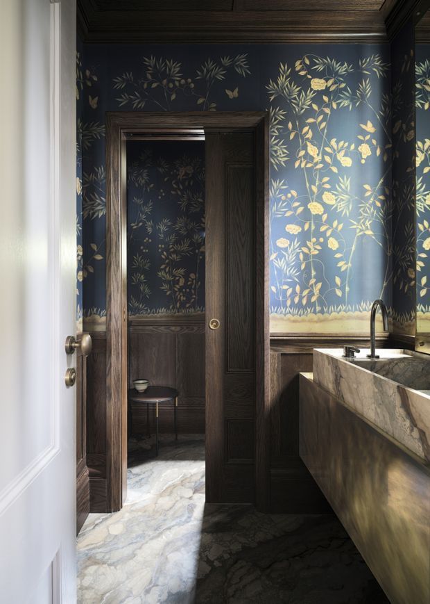 A richly detailed, dark-toned bathroom or powder room featuring deep navy botanical wallpaper with gold foliage, dark timber wall panelling and door joinery, a marble-clad vanity with an integrated basin and black curved tapware, and a pale veined stone floor. An open doorway reveals a small adjoining dark room with matching botanical walls and a stool.