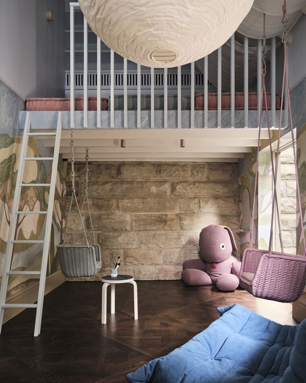 A playful, vertically arranged children's room with a lofted sleeping platform and white safety balustrade above a dark herringbone timber floor. The room features a ladder, hanging wicker-style swing seats, a large cream pendant light, a blue floor mattress or bedding, a plush mauve toy, patterned wall coverings, and a rustic exposed stone feature wall. Tall windows bring in natural light, while the layered levels create a compact, imaginative play-space atmosphere.