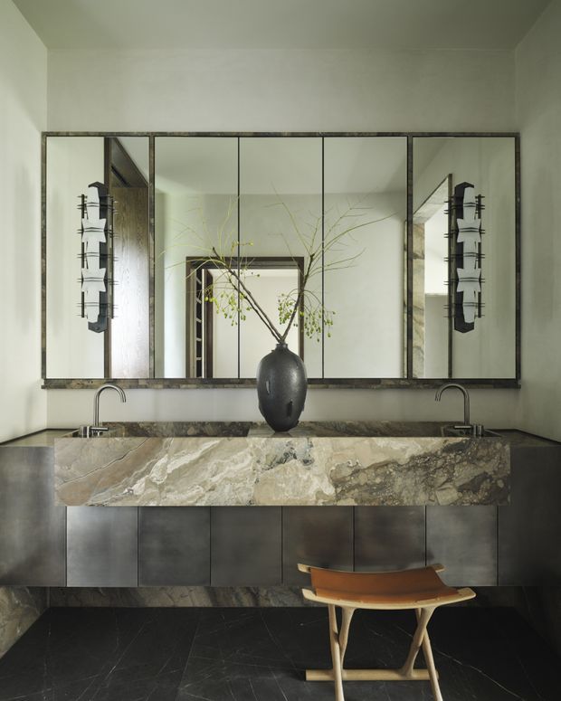 A sophisticated bathroom vanity with a long, heavily veined natural-stone benchtop and integrated twin basins, dark metal cabinetry, and slim black faucets. A large framed multi-panel mirror spans the wall above the vanity, reflecting a doorway and decorative branches. The room has dark stone flooring, pale plaster walls, sculptural wall sconces, and a warm wood-and-leather stool, creating a refined, moody interior.