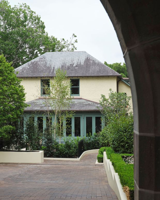 Exterior view of a two-storey residential building framed by a covered passage or entry structure. The home has a pale textured facade, dark grey hipped slate-like roof, teal-green timber-framed windows and doors, and a covered ground-floor verandah or porch. A landscaped courtyard and driveway use clipped hedges, raised rendered planters, mature trees, and brick paving.