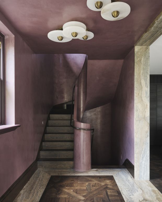 A dramatic interior stairway with dark timber treads, a sculptural curved balustrade and central newel finished in muted mauve plaster or stucco. The surrounding walls and ceiling are covered in a textured dusty-plum finish. A decorative geometric parquet floor occupies the landing, with a rough pale stone or plaster-clad opening on the right. Two vintage-style ceiling fixtures with frosted glass shades and brass details add a warm decorative accent. The space has a moody, luxurious and highly sculptural character.