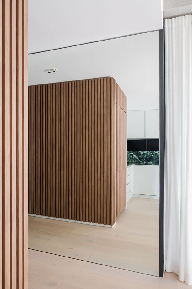 View through a doorway into a bright, minimalist contemporary interior. A large vertical timber slat feature wall or room divider dominates the foreground, with pale timber flooring, white walls and cabinetry, a dark benchtop or backsplash, and full-height white curtains. The space appears to be an open-plan kitchen area, with clean lines and concealed joinery.
