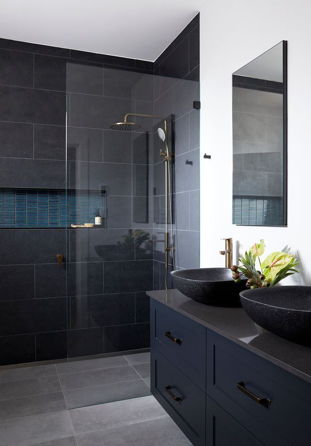 A sophisticated contemporary bathroom with a large walk-in shower enclosed by clear glass, dark charcoal wall tiles, matching grey floor tiles, and twin black vessel basins on a dark navy vanity. Brass-toned tapware and hardware provide warm accents, while a tall black-framed mirror and small plants complete the refined, moody scheme.