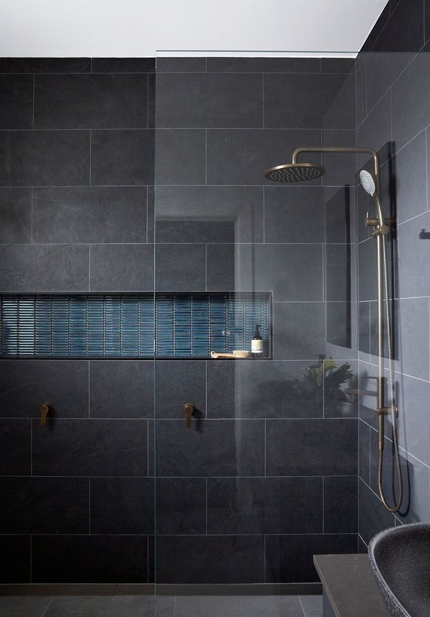 A dark, contemporary walk-in shower bathroom with large charcoal grey wall tiles, a frameless glass shower screen, and brushed brass rainfall and handheld shower fittings. A recessed horizontal niche with blue-grey mosaic tiles provides storage, while the edge of a dark freestanding bathtub and stone-like vanity surface are visible at the lower right.