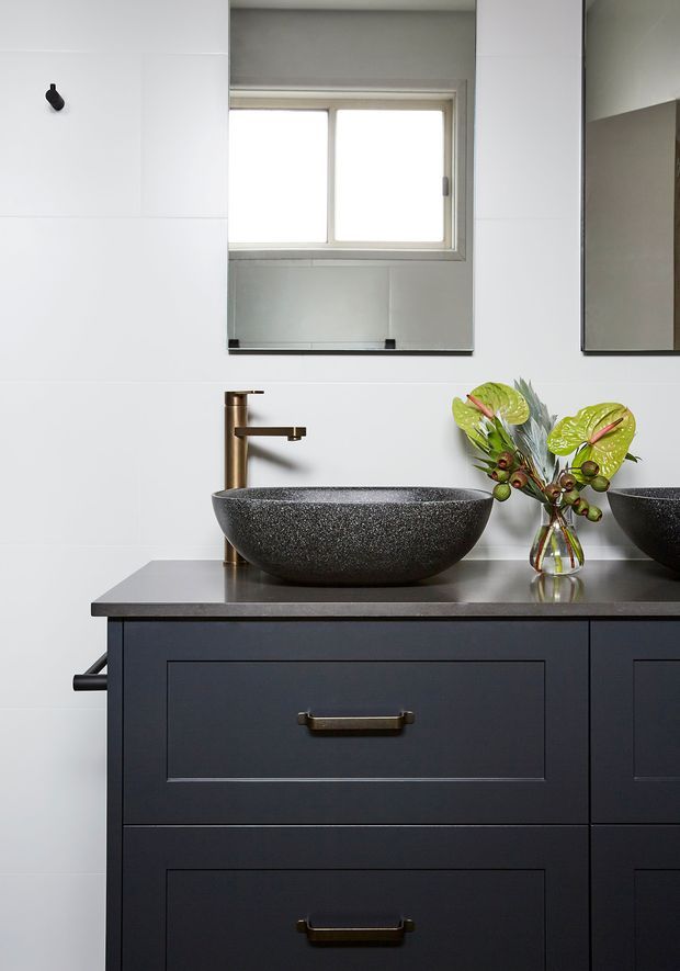 A refined bathroom vanity featuring a dark charcoal double-drawer cabinet, dark stone countertop, textured black vessel basin, and brushed brass wall-mounted faucet. White tiled walls, a large mirror, and a small window create a clean, contemporary setting, with greenery and flowers adding a natural accent.