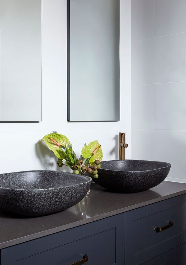 Close-up of a refined contemporary bathroom vanity with two large textured black vessel basins on a dark benchtop, dark blue-gray cabinetry with brass handles, pale tiled walls, frameless mirrors, and a small floral arrangement. The composition highlights the double vanity and contrasting dark fixtures against a light background.