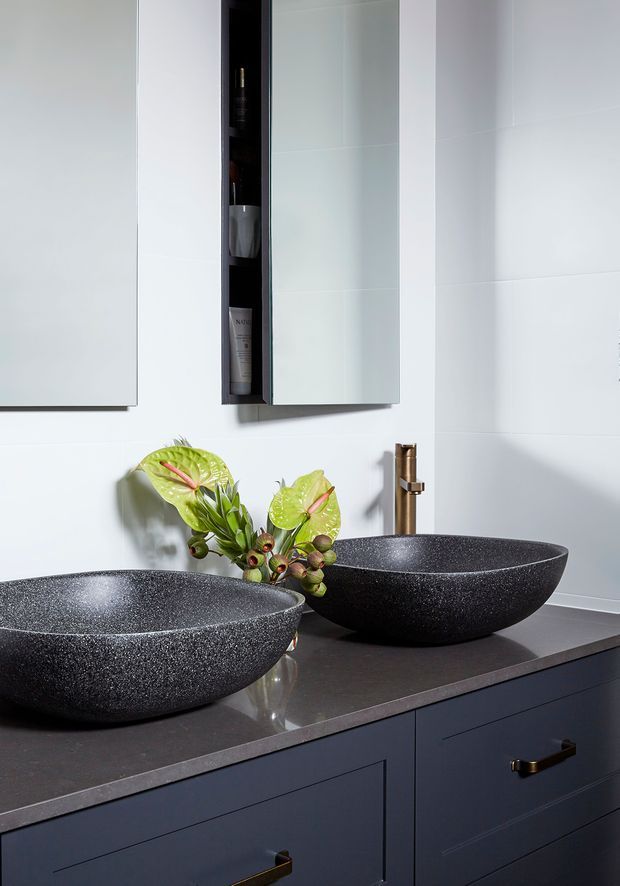 Contemporary bathroom vanity featuring two large round black textured vessel basins on a dark stone benchtop, dark blue-grey cabinetry with brass handles, a brass wall-mounted tap, and mirrored storage cabinets. Pale tiled walls and a small floral arrangement provide contrast.