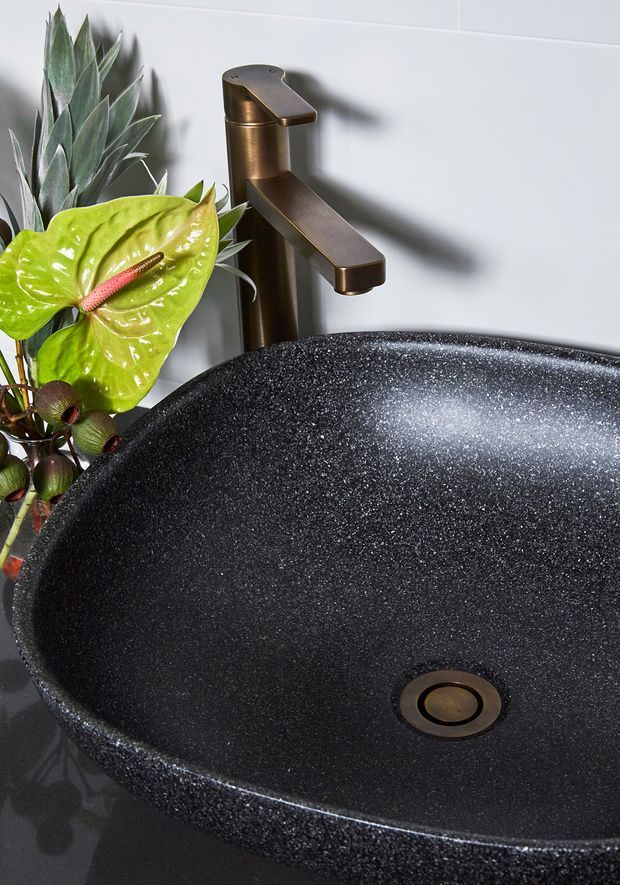 Close-up product-style image of a black, speckled countertop basin with a bronze-brown single-handle tap. The basin sits against a white tiled wall, with tropical foliage arranged beside it. The composition highlights the basin texture, rounded form, and coordinating tapware.