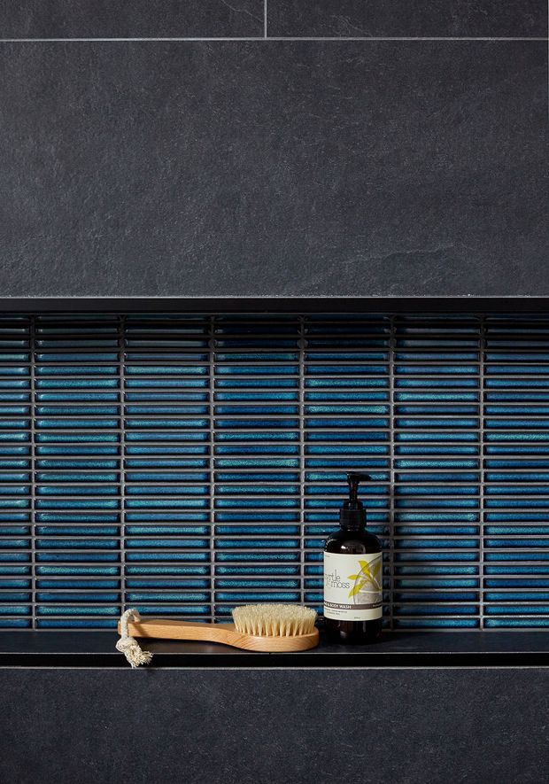 Close-up detail of a dark tiled wall with a recessed shelf lined in glossy, horizontally arranged blue-black mosaic tiles. A wooden body brush and dark pump bottle sit on the shelf, creating a refined, moody bathroom finish detail.
