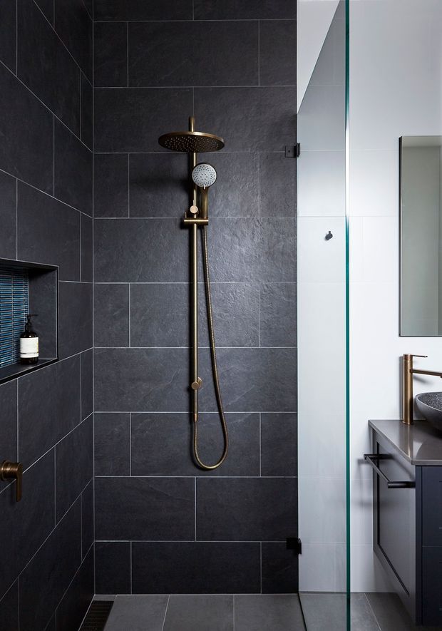 A contemporary bathroom shower area with large dark charcoal wall and floor tiles, a bronze rain shower and handheld fixture, recessed wall niche, and frameless clear glass shower screen. A dark vanity with a grey benchtop, bronze tapware, vessel basin, and wall mirror is partially visible at right.