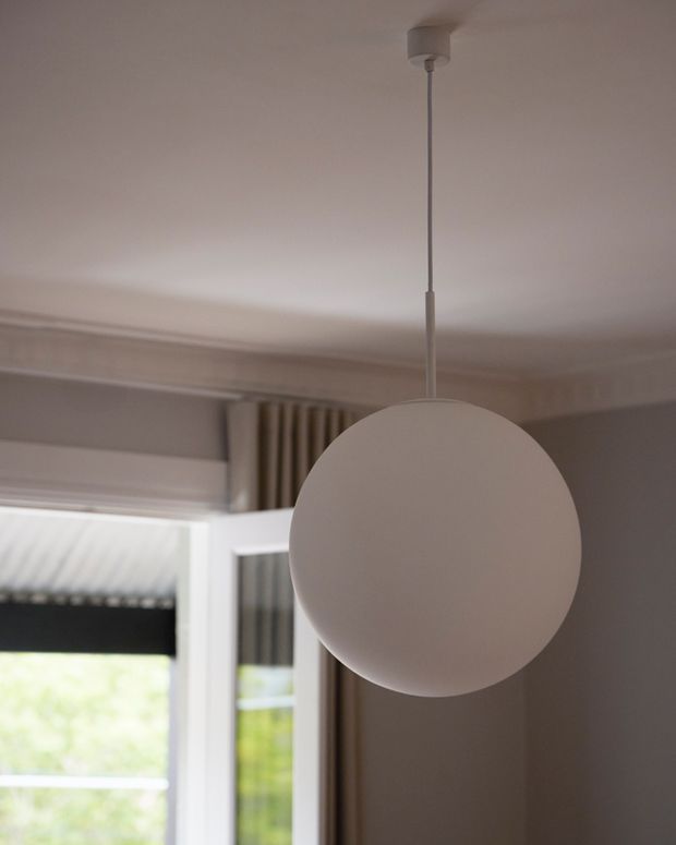 Close-up interior view of a simple spherical pendant light suspended from a white ceiling. The fixture hangs in front of a window with vertical curtains and roller blinds, with restrained neutral finishes and traditional ceiling cornice detailing visible in the background.