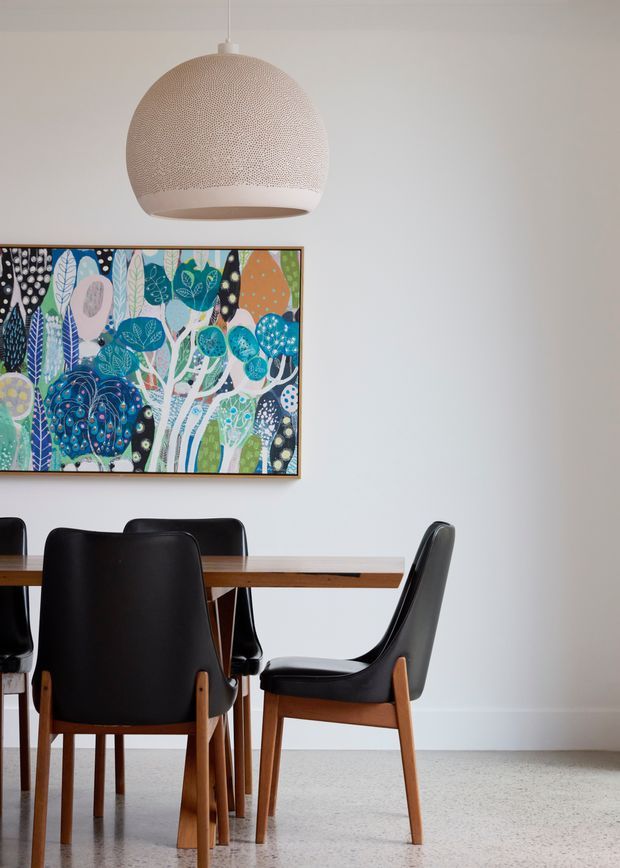 A bright dining area with a rectangular timber dining table surrounded by black upholstered mid-century-style dining chairs. A colourful botanical artwork hangs on the white wall, complemented by a large textured globe pendant light. The room has a pale terrazzo-like floor and a restrained, contemporary palette with vivid artwork accents.
