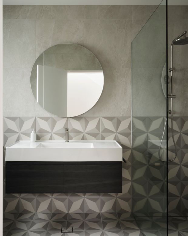 Contemporary bathroom featuring a floating dark wood vanity with a white integrated basin, round wall mirror, chrome tapware, and a frameless glass shower enclosure. Large-format pale stone-look wall tiles are paired with geometric three-dimensional patterned tiles across the lower walls and floor, creating a refined monochromatic feature surface.