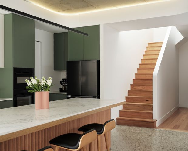 Contemporary open-plan kitchen with deep sage-green cabinetry, black appliances, a large white marble-look island with a timber fluted base, and two curved black-and-timber bar stools. A timber staircase with a white sculptural balustrade rises beside the kitchen, while an exposed concrete ceiling and integrated linear lighting add a refined modern character.