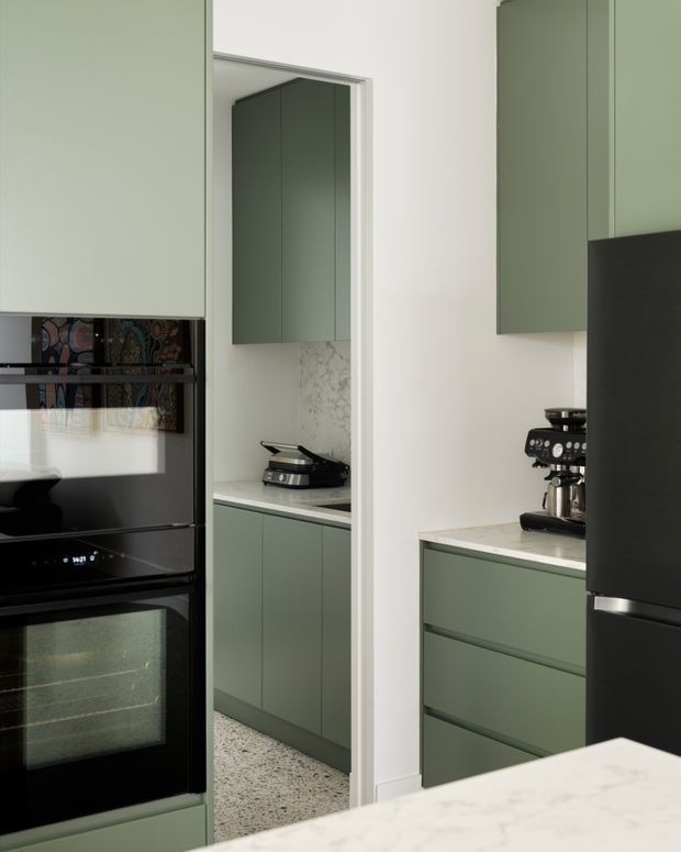 A compact contemporary kitchen in muted sage-green cabinetry with integrated black appliances, a light stone benchtop, and terrazzo flooring. A mirrored opening reveals a pantry or secondary preparation area with additional cabinetry, a marble-patterned splashback, and a sandwich press. A coffee machine sits on the main benchtop, while a white island or counter occupies the foreground.