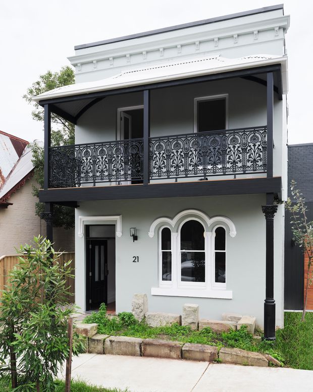 Street-facing view of a narrow two-storey heritage-style residence with a pale rendered facade, ornate black lacework balcony, projecting upper-level verandah, decorative white cornice detailing, classical columns, arched multi-pane window, and a small landscaped front garden.