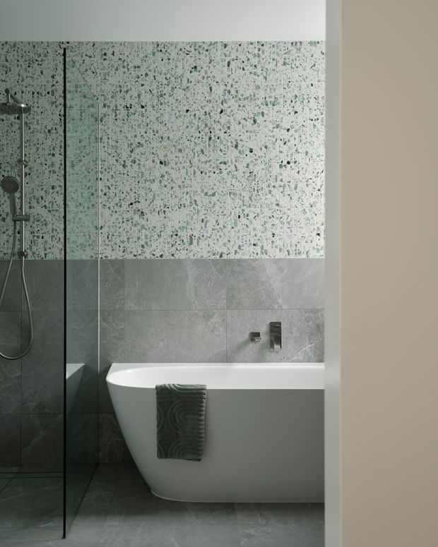 Contemporary bathroom featuring a freestanding white bathtub, walk-in shower with clear glass screen, grey stone-look floor and wall tiles, and a speckled terrazzo-style feature tile wall. Brushed metal shower and tap fittings add understated contrast.
