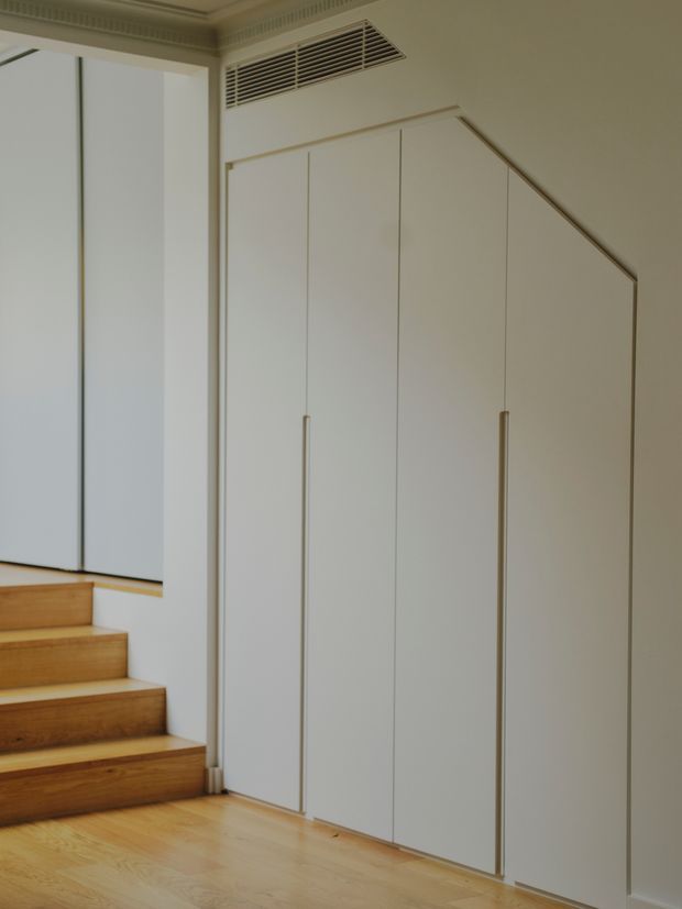A refined interior hallway and stair landing featuring a tall, custom built-in wardrobe or storage wall with minimalist white panelled doors and slim vertical handles. Warm timber stairs and flooring contrast with the pale cabinetry and walls. Decorative ceiling moulding, a concealed air-conditioning grille, and a sharply angled upper cabinet edge are visible.