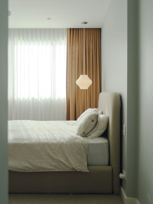 A softly lit, compact bedroom viewed from the doorway, featuring a neatly made upholstered bed with a tall rounded headboard, white bedding and pillows, floor-to-ceiling tan curtains, sheer window drapes, and a sculptural pendant light. The room has pale walls, a light carpeted floor, and a calm, refined contemporary feel.