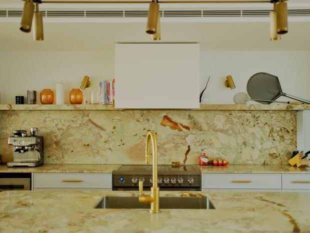 A contemporary kitchen featuring a large island with an integrated stainless-steel sink and prominent brass gooseneck tap. The cabinetry is light grey with slim brass handles, while the benchtops and full-height splashback are clad in richly veined beige and cream natural stone. Open shelving displays books and decorative objects, with brass track lighting and a built-in cooktop along the rear wall.