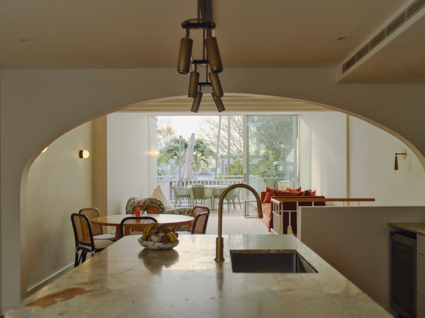 Warm contemporary open-plan kitchen looking through a broad curved archway toward a dining and living area with floor-to-ceiling glazing. The foreground features a large pale stone island with an integrated sink and brass-toned tap, dark cabinetry, pendant lighting, and a dining table beyond. Outdoor greenery is visible through the glazing.