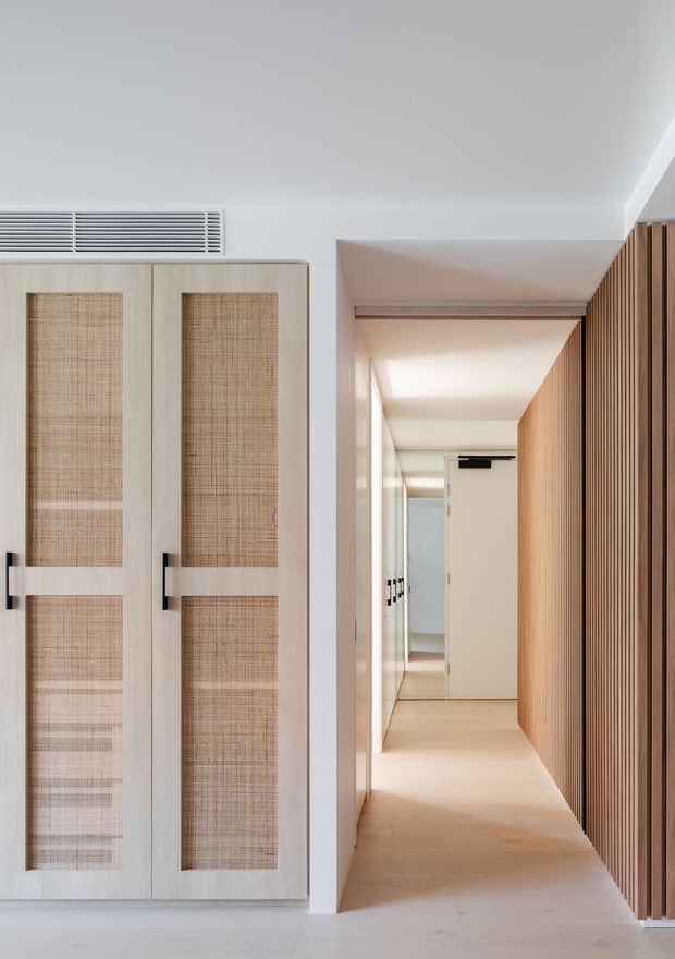 A bright, refined residential hallway with pale timber flooring and a long corridor leading to a closed door. The left side features tall built-in cabinetry with light timber doors and woven natural-fibre inset panels, while the right wall is clad in vertical timber battens. White walls and ceiling, concealed doorways, and integrated linear air-conditioning grilles create a clean, contemporary interior with strong warm wood detailing.