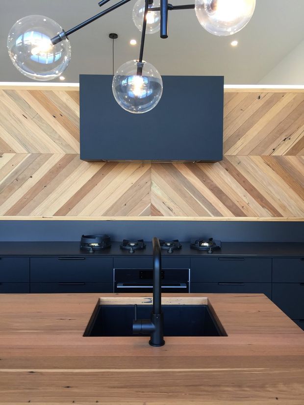A contemporary kitchen viewed straight on, featuring deep navy cabinetry, a timber island benchtop with an integrated dark sink and black tap, and a dramatic herringbone timber splashback. A matching navy rangehood is mounted above the cooktop, while clear globe pendant lights hang overhead.