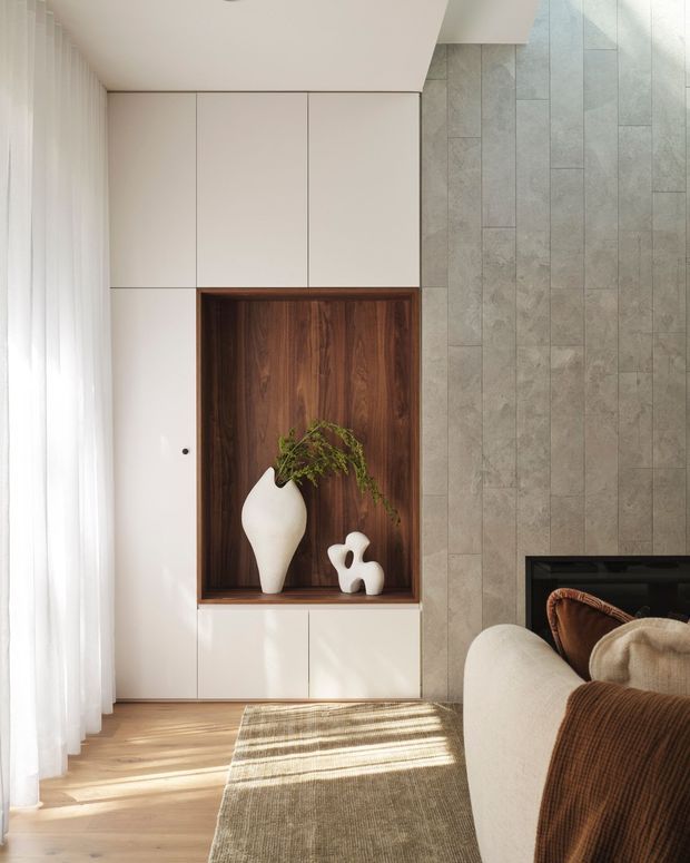 A softly lit contemporary living room with floor-to-ceiling sheer curtains, pale timber flooring, a built-in white cabinetry wall, and a recessed dark timber display niche holding sculptural ceramics and greenery. A large-format grey stone-look tiled feature wall surrounds a dark fireplace, with a cream sofa, brown textured throw, and neutral woven rug in the foreground.