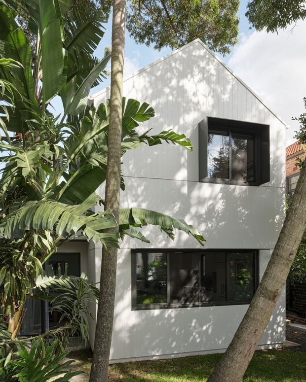 Exterior view of a contemporary two-storey house extension or residence, surrounded by dense tropical and subtropical planting. The facade is clad in light grey-white vertical panels, with dark recessed window frames and large glazed openings. A steep gable roof is visible, while mature trees cast dappled shadows across the building.