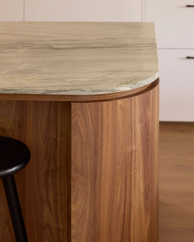 Close-up detail of a rounded kitchen island or peninsula with a warm timber veneer base and a pale, veined natural stone benchtop. A dark timber or black stool is partially visible, with white cabinetry and timber flooring in the background.