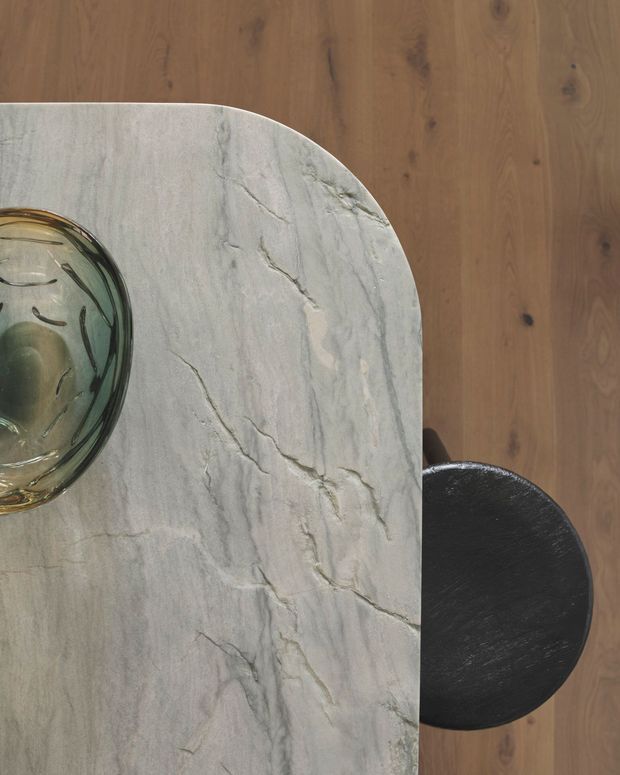Top-down close-up of a rounded rectangular stone kitchen island or table with prominent grey veining, paired with a dark round stool and a translucent glass bowl. Warm timber flooring surrounds the fixed stone surface, creating a restrained, tactile contemporary interior detail.