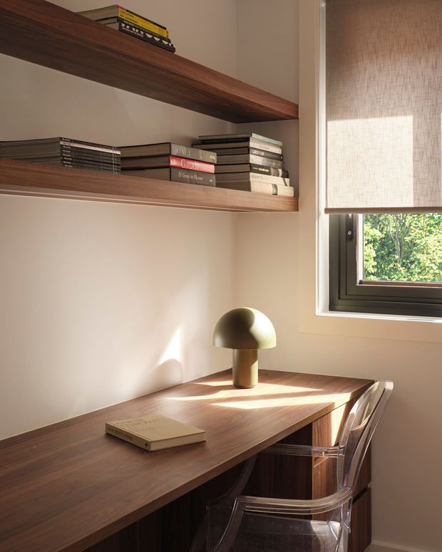 A compact, warm home office nook with a built-in dark timber desk and matching open shelving filled with books. A small dome-shaped table lamp, transparent acrylic chair, roller blind, and black-framed window complete the softly sunlit space.