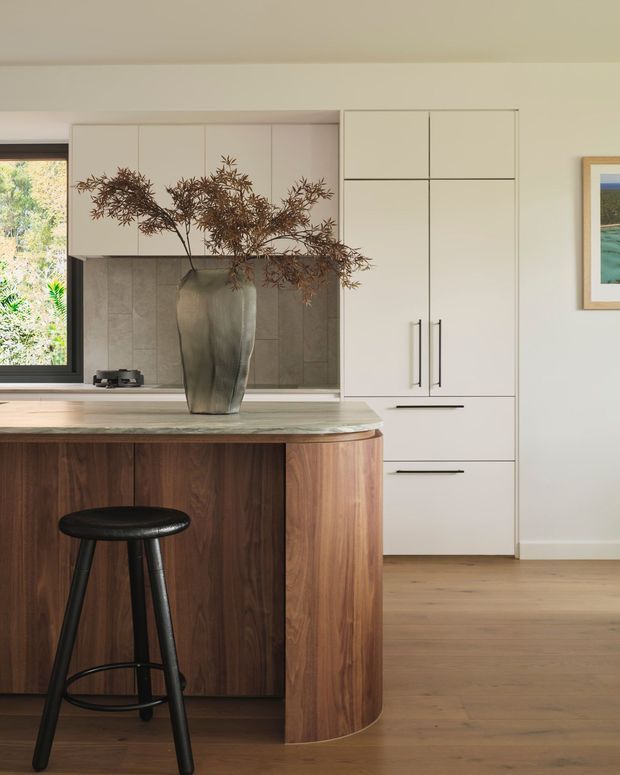 A warm, refined kitchen interior featuring a central island with rich walnut cabinetry and a rounded end, topped with a pale stone benchtop. A black bar stool sits in front of the island. Behind it are white full-height cabinets, a grey tiled splashback, integrated appliances, and a large window overlooking greenery. Timber flooring and a framed artwork complete the calm, contemporary space.