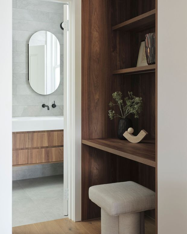 Interior view of a refined bathroom and adjacent built-in dark timber shelving. The bathroom contains a freestanding-style vanity with timber drawers, pale grey wall and floor tiles, a rounded wall mirror, and matte black plumbing fixtures. In the foreground, the fixed shelving displays decor and includes a padded upholstered stool, creating a warm, contemporary transition between spaces.