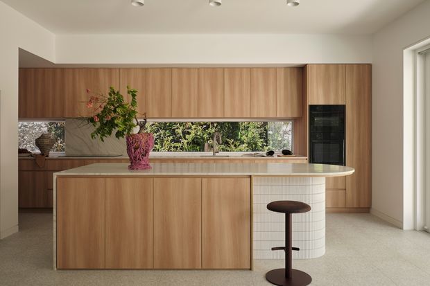 Warm contemporary kitchen with extensive mid-tone timber cabinetry, a large stone island with a curved tiled end, integrated appliances, and a full-width window splashback overlooking greenery. The space has a restrained, refined palette with concealed storage and recessed ceiling lights.