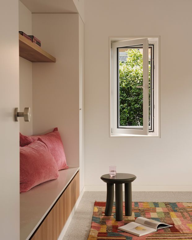 A compact, warmly styled bedroom nook with a built-in window seat featuring pink cushions and integrated wood-front cabinetry. A floating timber shelf with books occupies the adjacent alcove. The room has pale warm walls, a small dark round side table, a colourful patterned rug, and a white-framed casement window overlooking greenery.