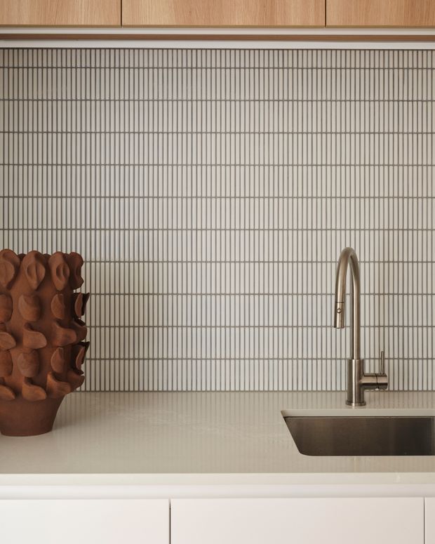 Close-up of a refined contemporary kitchen detail featuring pale vertically grooved wall tile, warm light-oak overhead cabinetry, a pale stone or solid-surface benchtop, white lower cabinetry, and a brushed metal gooseneck mixer beside an undermount stainless-steel sink. A sculptural brown vessel sits on the counter.