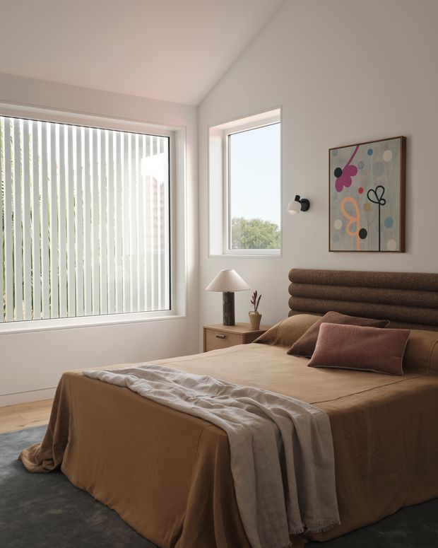 A warm, softly lit contemporary bedroom with a large bed dressed in earthy brown bedding, layered taupe and rust cushions, and a textured upholstered headboard. A timber bedside table holds a lamp and small plant, while abstract artwork and a wall sconce decorate the room. Large windows with vertical blinds provide daylight and views of greenery.