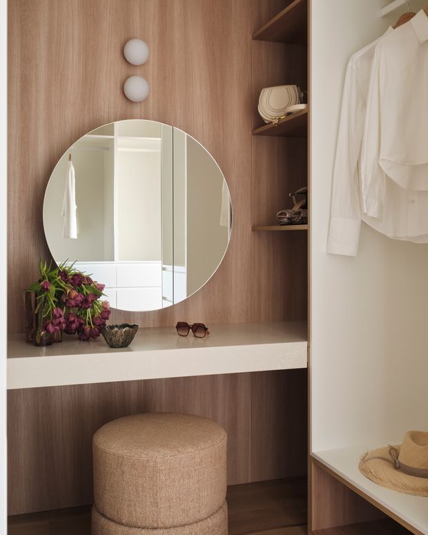 A refined walk-in wardrobe or dressing nook with dark timber veneer joinery, open shelving, a white stone-look dressing counter, large round mirror, integrated globe wall lights, hanging white clothing, accessories, and a textured upholstered stool. The space has warm, curated styling and built-in storage.