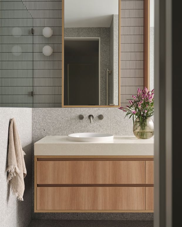 A refined contemporary bathroom vanity featuring a wall-mounted natural timber drawer unit, pale stone benchtop with a white vessel basin, wall-mounted tapware, and a large mirror. Grey terrazzo-like wall and floor surfaces contrast with a dark vertical tiled feature wall, while a vase of flowers and a hanging towel add softness.