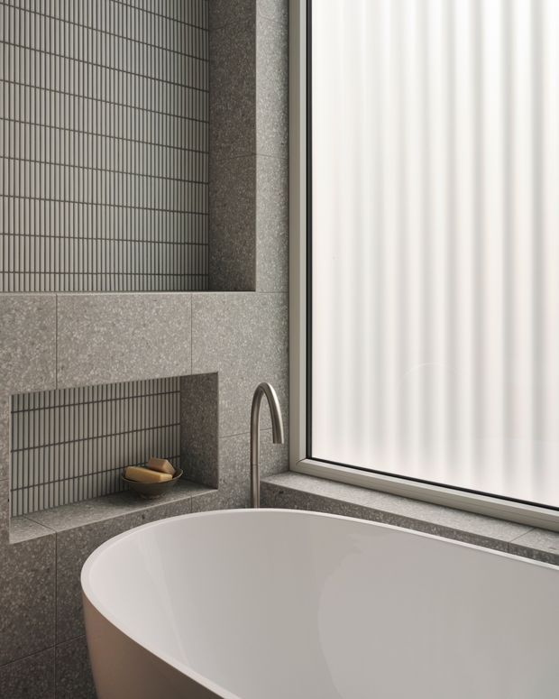 Close-up view of a refined contemporary bathroom featuring a freestanding white bathtub beside a large frosted window. The walls and recessed niche are finished in grey stone-look surfaces with narrow vertically oriented dark grey tiles, and a curved brushed metal wall-mounted tap sits above the bath edge. Soft diffused daylight creates a calm, minimalist atmosphere.