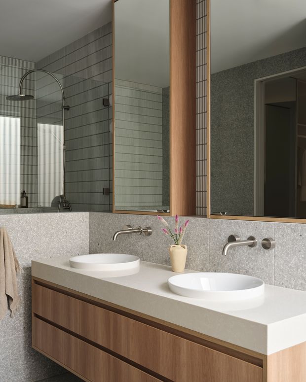Contemporary double-basin bathroom with a long timber vanity, white vessel basins, pale solid-surface benchtop, grey terrazzo-like wall panelling, and vertically gridded grey tile walls. A large mirror, wall-mounted brushed metal taps, and an arched shower fixture contribute to the refined, minimalist composition.